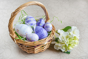 Easter wicker basket with colored eggs and a white flower on white board.