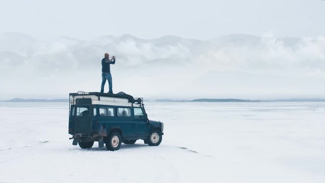 WIDE Caucasian male standing on the roof of old 4x4 off-road vehicle, taking pictures and enjoying the view of mountains over large lake. 4K UHD 60 FPS RAW edited footage