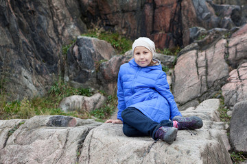 Young Caucasian girl in blue jacket sitting on a rock