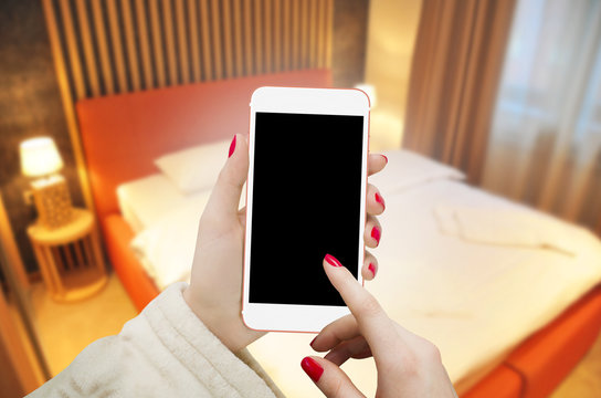 White smartphone in female hands with isolated blank screen. Free space for text. Blurred hotel bedroom in background. 