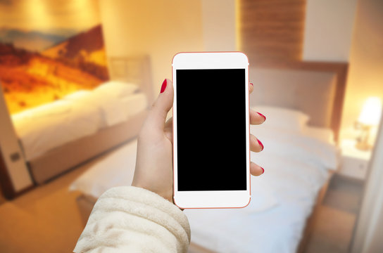 Women holding white phone in hand with bathrobe sleeve. Isolated black blank screen for text input. Bedroom in background.