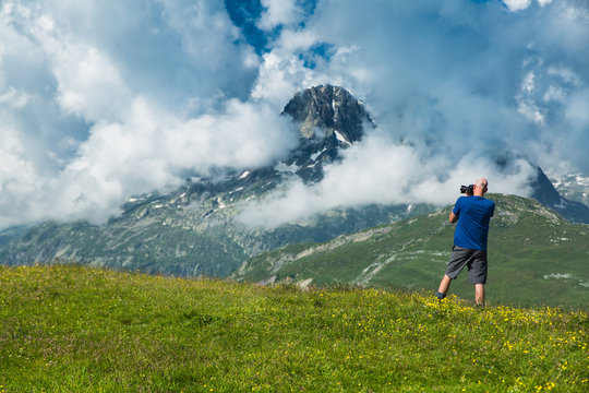A tall bald man takes photos of the mountain valley and snow covered peaks of the French alps