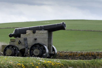 Orkneys, Scotland - June 5, 2012: A black historic cannon stands in the wider area around Skara Brae Neolithic settlement standing in green meadow with white and yellow wild flowers. . White-blue sky.