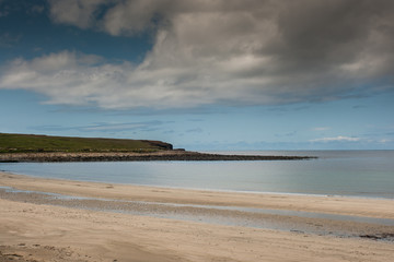 Orkneys, Scotland - June 5, 2012: Skara Brae Neolithic Settlement. The light gray Atlantic Ocean with a stretch of the south shore protruding in the water. Sand and a dark cloudy blue sky.