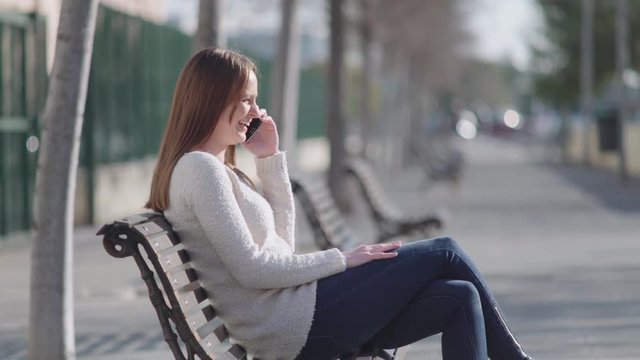 Woman sitting in a bench at the park. She talks by cell phone and laughs. The camera moves around.