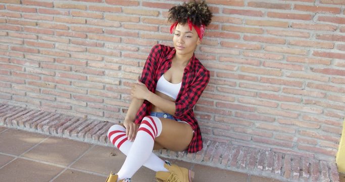 Fashionable sexy african american girl sitting on urban street. She wearing skimpy denim jeans  high knee socks and checkered red shirt. She leaning against brick wall.