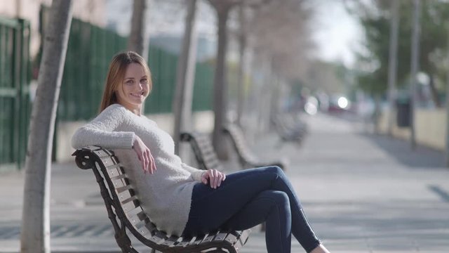 Woman sitting in a bench at the park. She looks at camera and smiles.