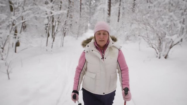 Energetic woman in warm white waistcoat and black trousers doing active nordic walking exercising in the forest. Female athlete stepping quickly with ski poles on the path in winter nature outdoors.