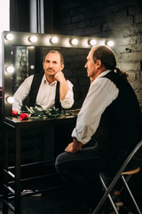 Handsome middle aged man is dressed in a white shirt and a black vest, holding a red rose flower, bachelor. Black background.