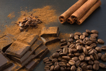 Black chocolate pieces, cinnamon and coffee beans, chocolate slabs spilled from grated chocolate powder on black slate dish. Close up.