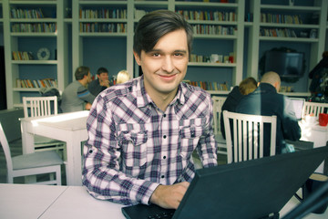 Young smiling man in checkered shirt working in cafe. Book shelves on background. Freelance concept. 