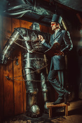 Young bearded man in cylinder hat is reading manual book while standing near huge robot.