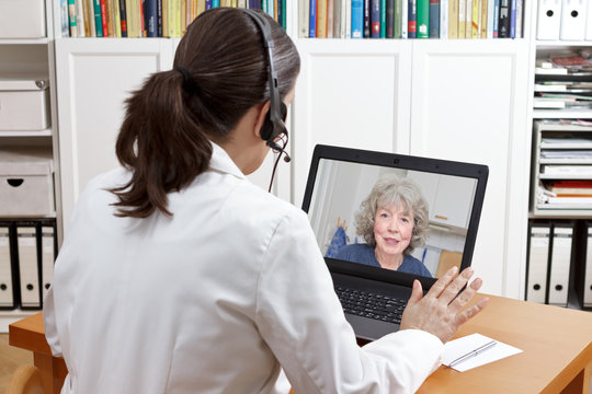 Female doctor of geriatrics in her surgery office with headset in front of her laptop talking via video call with an old patient about her prescribed drugs