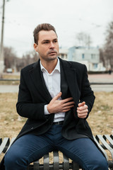 Fototapeta premium the man in a black coat and a suit sits on a bench. He looks for something in a pocket. He seems something has left at home, he has a confused look.