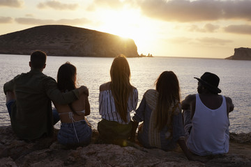 Fototapeta na wymiar Rear View Of Friends Sitting On Cliff Watching Sunset
