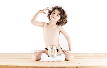 Cute curly-haired boy in shorts sitting in a "position of the Hero," his hand on his head. Before him is a wooden mug and towel. White background.