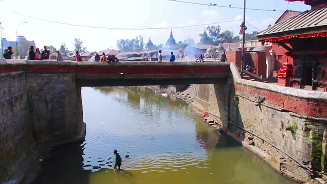 People crossing and walking on a stone bridge over a river in rural Asia with the sky and sunlight reflecting in the water where a child is wading and playing