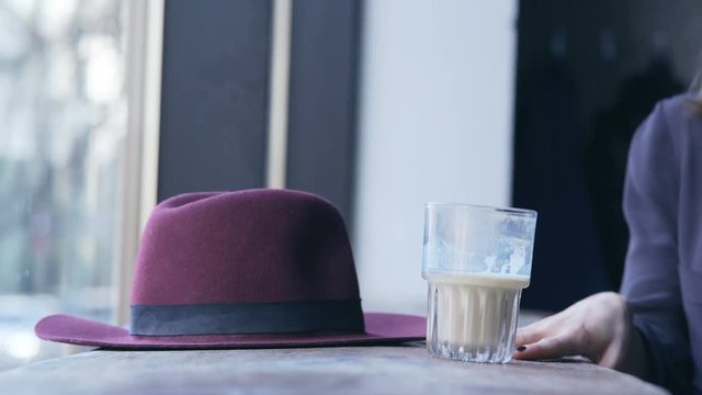 Smiling young girl sits and putting her hat and drink on the table in cafe