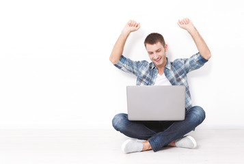 Handsome young man sitting on the floor near the wall, celebrating success with arms raised while looking at his laptop screen