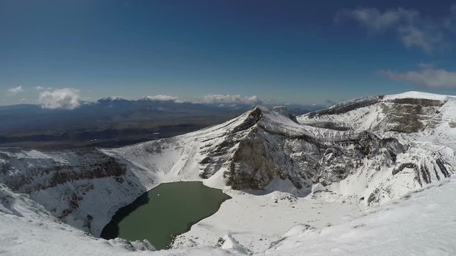 Volcanic landscape of Kamchatka Peninsula: beautiful view of snowbound craters and crater lake of active Gorely Volcano on a sunny day with dark blue sky.