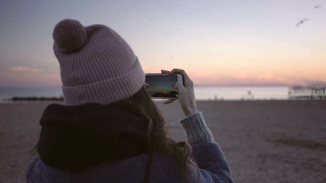 Back view of Woman standing on the beach making photo on the smartphone.