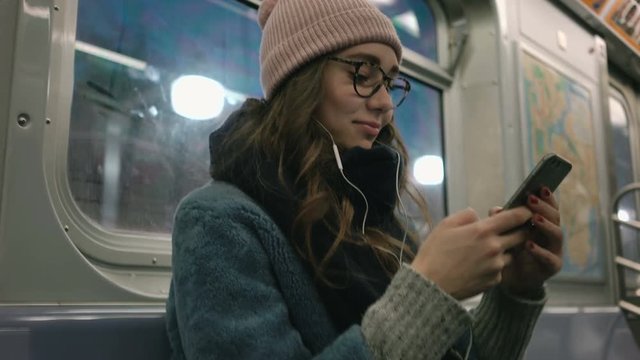 View from below of Woman in warm clothes and eyeglasses using smartphone and sitting in metro carriage