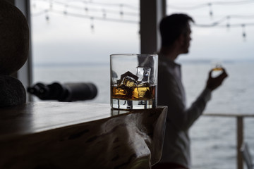 Scotch  on the Rocks with man in background enjoying window view.