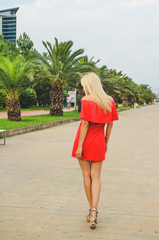 Girl posing in the red dress on the quay. Pretty blonde woman surrounded by tropical palms near sea Rear view walking girl