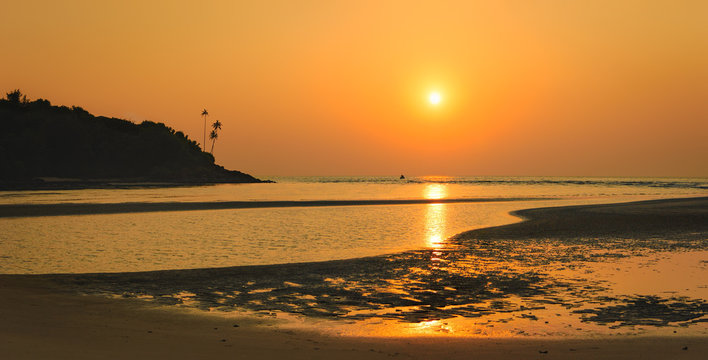 Bright tropical sunset at low tide on the Mobor Beach, South Goa, India. Beautiful seascape with silhouettes of palm trees on the mountain and fishing boats on the horizon