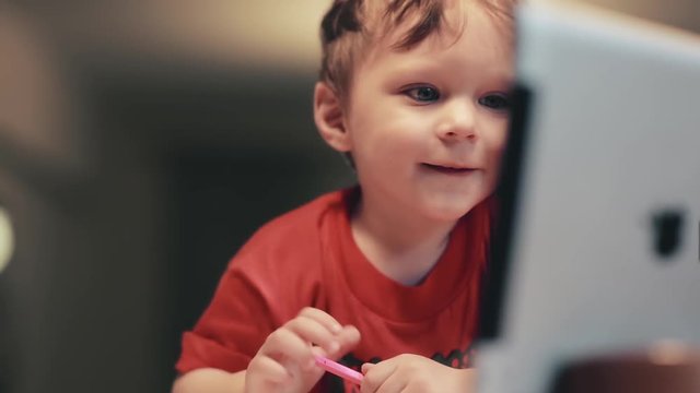Cute little boy uses a digital tablet, smiling and drawing with the pink color pencil.