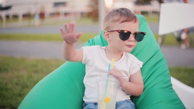 Funny little boy in a big sunglasses sitting in bag chair at the park in summer. Holding a glass and dancing like rapper