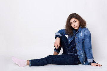 Fototapeta premium A cute girl wearing stylish jean clothes sitting on the floor indoors isolated over white background looking into camera with her charming dark eyes relaxing at home. Relaxed young woman in jeans