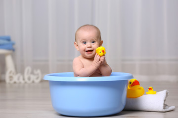Cute baby playing with rubber duck while sitting in plastic basin at home