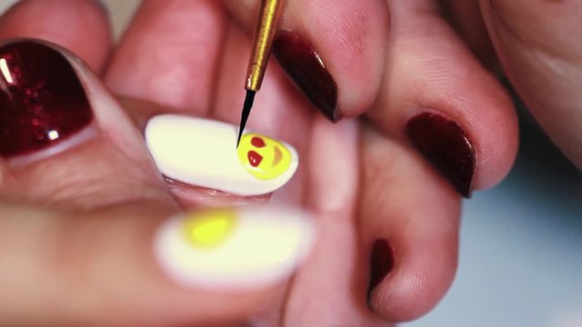 Manicure procedure female hand painting yellow emoji on white finger nail polish, close up