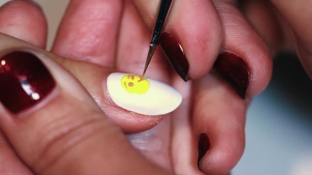 Manicure procedure woman hand draws yellow emoticon on white finger nail varnish, close up