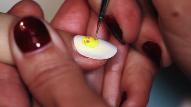 Manicure procedure female hand paints yellow emoticon on white finger nail polish, close up