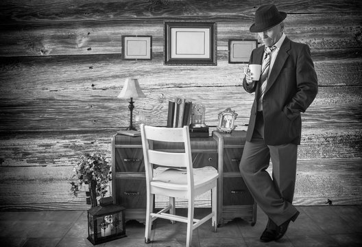 horizontal black and white image of a caucasian man in a  business suit and hat standing by his desk holding a cup of coffee.