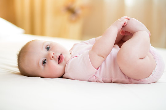 Concept of healthy child. Cute baby lying on her back on bed in room, holding legs with her hands.