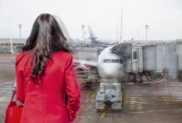 Woman in red dress waiting for flight in modern airport terminal building, watching airplanes in window on rainy day