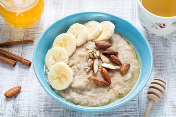 Oatmeal porridge bowl with banana, sliced almonds, sunflower seeds and honey. Healthy nutritious breakfast with cup of green tea. Closeup