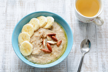 Oatmeal porridge bowl with banana, sliced almonds, sunflower seeds and honey. Healthy nutritious breakfast with cup of green tea. Top view