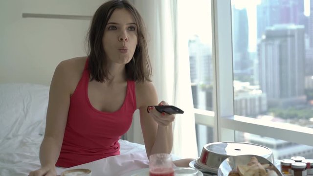 Young woman watching TV and eating breakfast on bed at home
