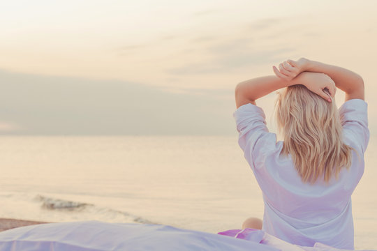 blond woman wakes up on the beach, sitting in a bed of silk on the sand, wearing a man's shirt, greets the dawn