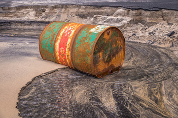 Rusty barrel oil on a partly black coloured beach illustrates the pollution of environment by oil spills