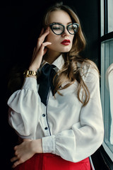 Indoor portrait of a young beautiful fashionable business woman posing in loft interior. Model looking through window. Lady wearing stylish clothes. Female fashion concept. Dark background. Waist up