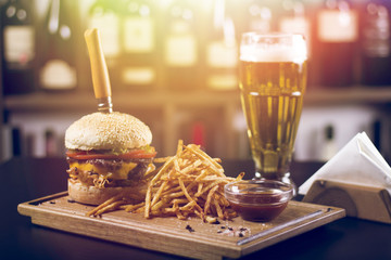 Close up of a delicious cheeseburger, tomatoes, and french fries, beer in the background. Beautifully served meal at the restaurant. Lens flare in the background.