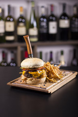 Close up of a delicious cheeseburger, tomatoes, and french fries. Pickles and cheese dripping off the side. Knife stabbed through a burger on a kitchen table. Wine bottles in the background.