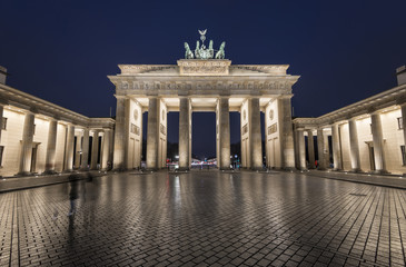 Fototapeta premium oświetlona Brama Brandenburska (Brandenburger Tor) odbijająca się na mokrym bruku o niebieskiej godzinie wieczorem, Berlin Mitte, Niemcy, Europa