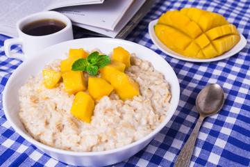 Oat porridge with fresh mango in the white bowl with coffee and book