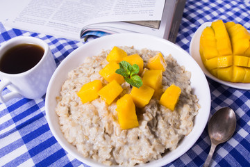 Oat porridge with fresh mango in the white bowl with coffee and book